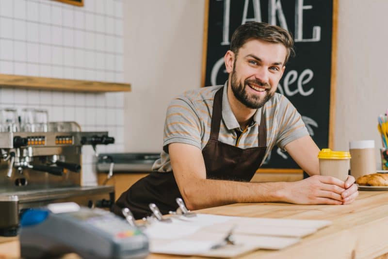 Friendly male barista smiling at the café counter, holding a takeaway coffee cup, with payment terminal and espresso machine visible.
