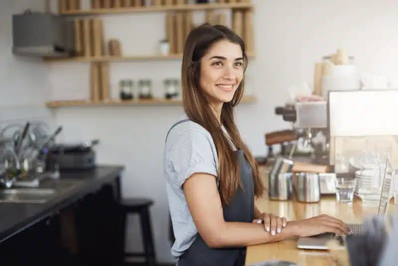 Smiling female café owner wearing an apron standing at the counter using a laptop in a modern coffee shop setting.