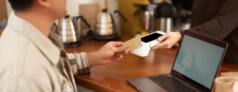 A customer makes a contactless payment with a smartphone at a coffee shop counter, tapping against a wireless card reader held by a barista.