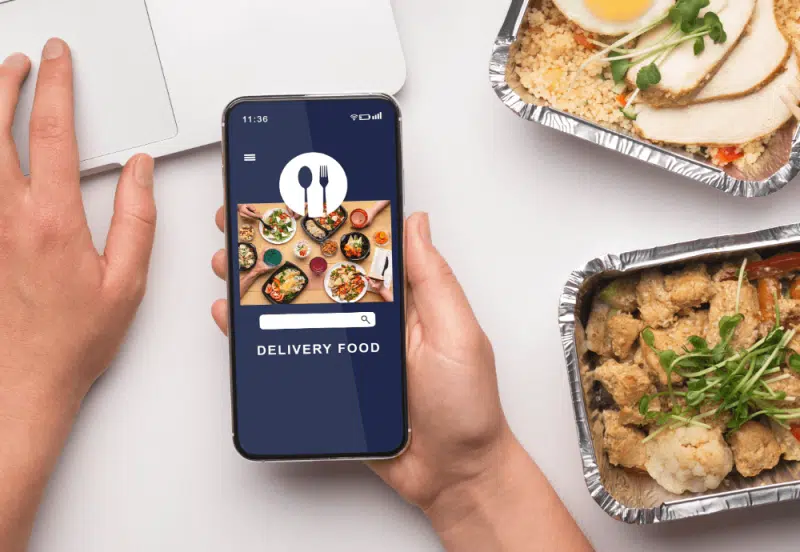 Hand holding a smartphone with food delivery app open alongside takeaway meal containers.