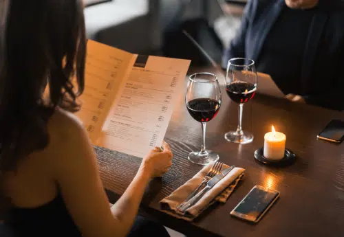 A couple sitting at a dimly lit restaurant table with glasses of red wine, looking at menus by candlelight.