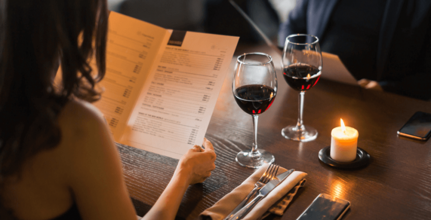 A couple sitting at a dimly lit restaurant table with glasses of red wine, looking at menus by candlelight.